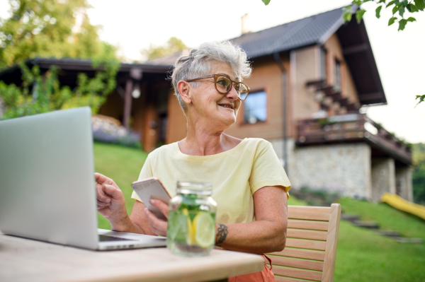 Happy senior woman with laptop, smartphone working outdoors in garden, home office concept.