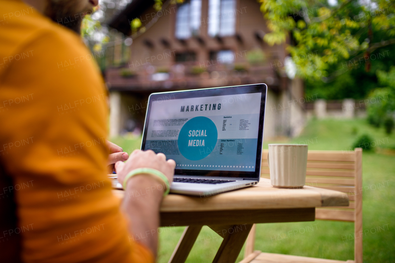 An unrecognizable man with laptop working outdoors in garden, home office concept.