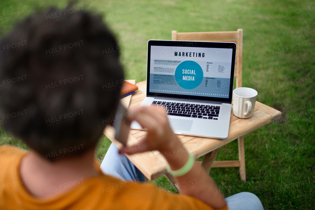 Rear view of mature man with laptop and smartphone working outdoors in garden, home office concept.