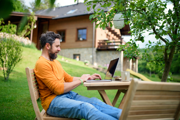 Side view of mature man with laptop working outdoors in garden, home office concept.