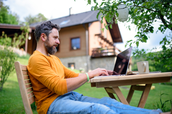 Side view of mature man with laptop working outdoors in garden, home office concept.