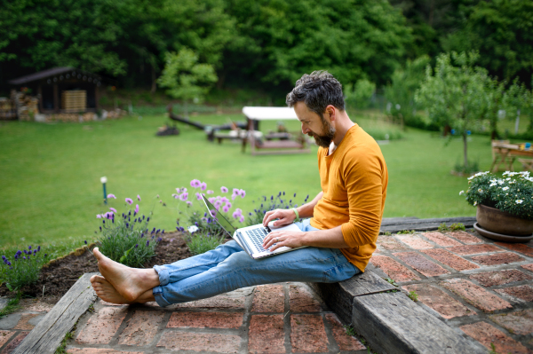 Portrait of mature man with laptop working outdoors in garden, green home office concept.