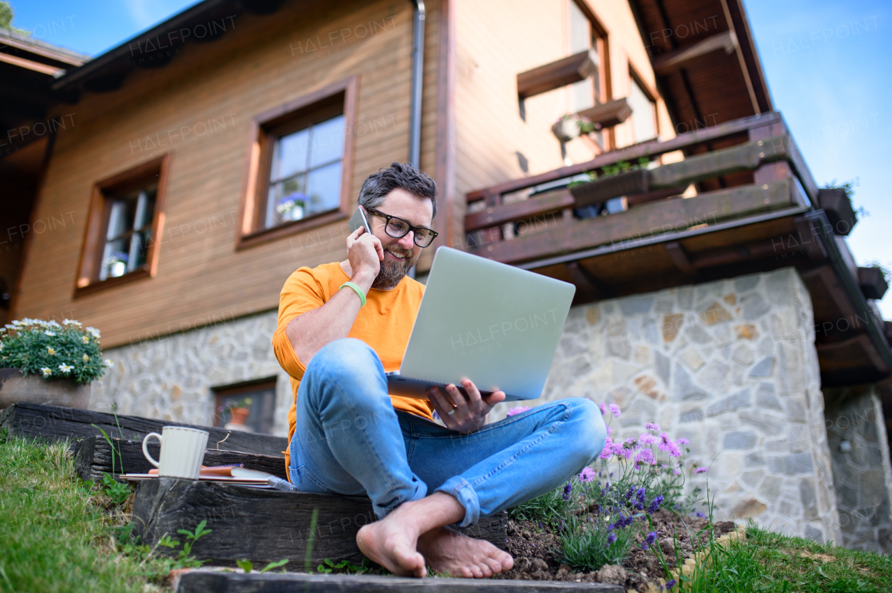 Portrait of mature man with laptop and smartphone working outdoors in garden, home office concept.