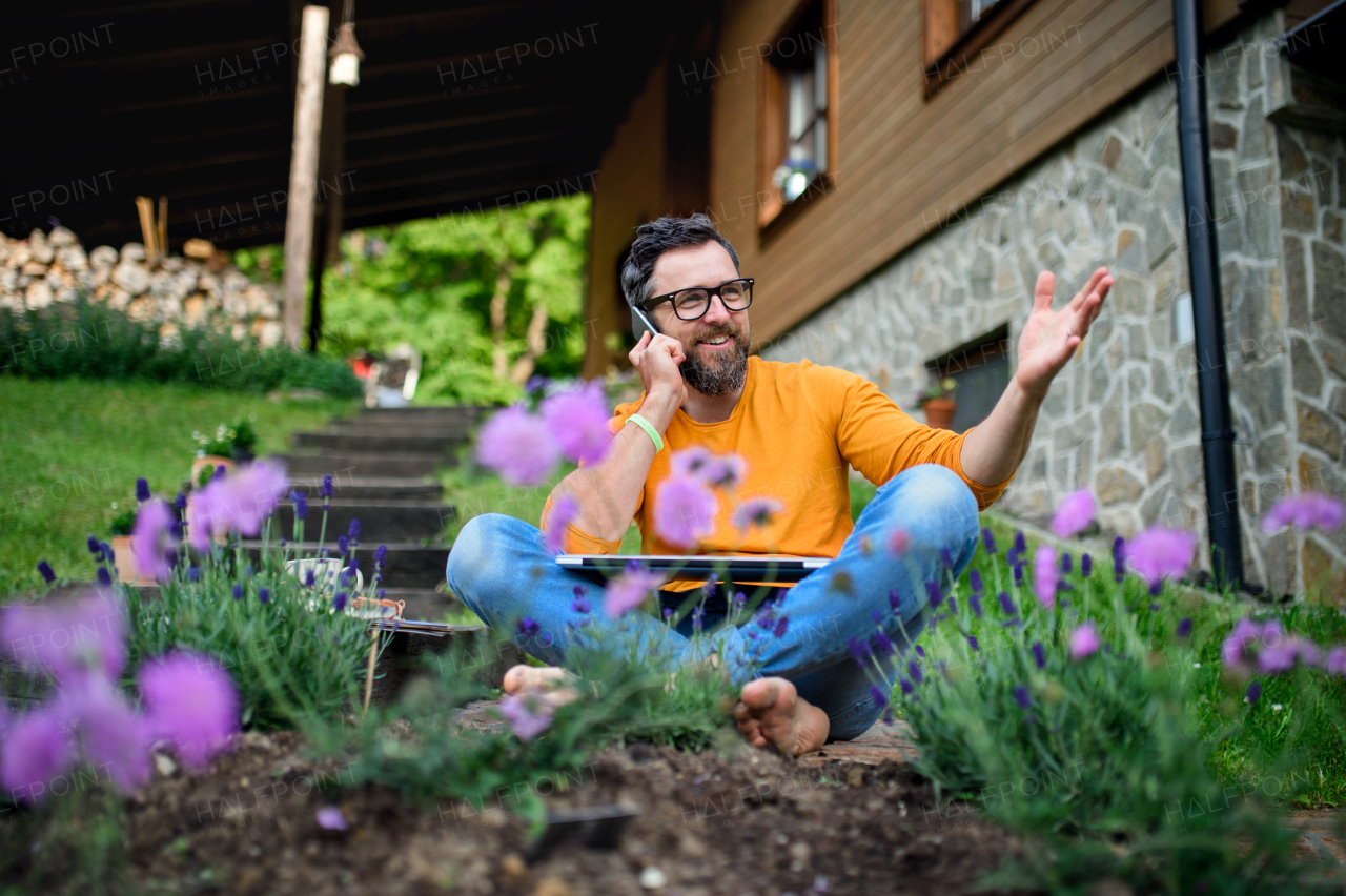 Portrait of mature man with laptop and smartphone working outdoors in garden, home office concept.