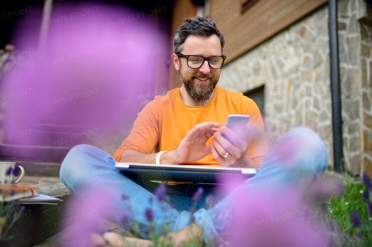 Portrait of mature man with smartphone working outdoors in garden, green home office concept.