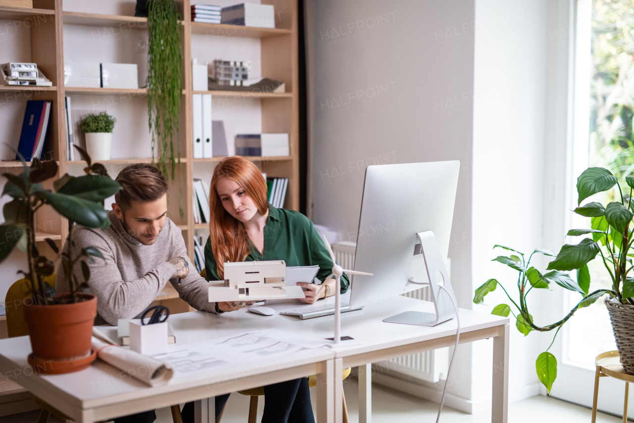 Young architects with model of house sitting at the desk indoors in office, working.