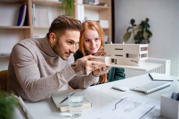 Young architects with model of house sitting at the desk indoors in office, working.