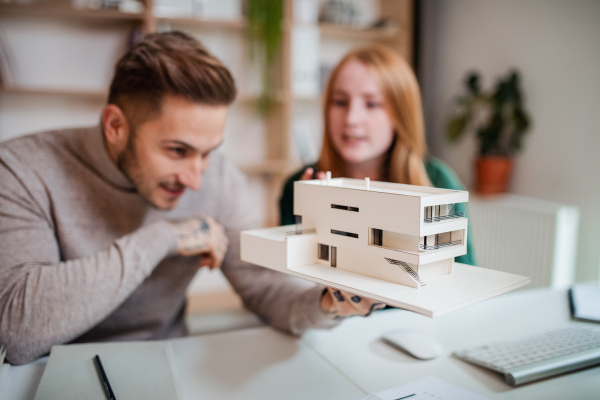 Young architects with model of house sitting at the desk indoors in office, working.