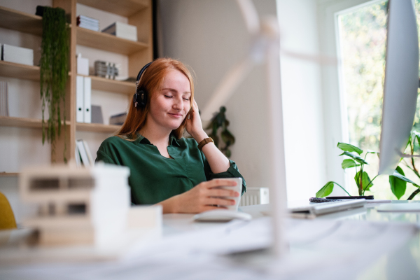 Young businesswoman with headphones sitting at the desk indoors in office, listening to music.