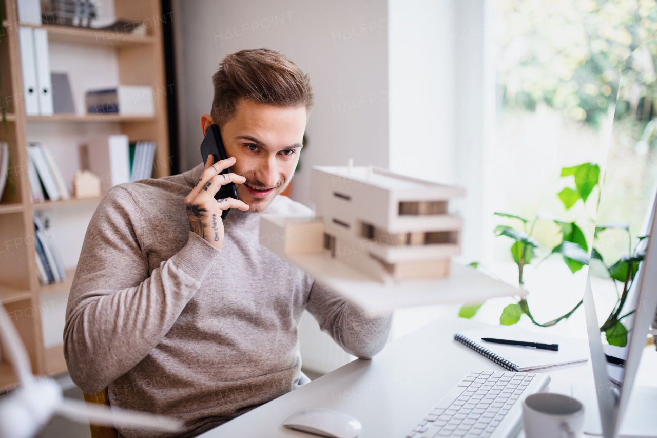 A young architect sitting at the desk indoors in office, using smartphone.