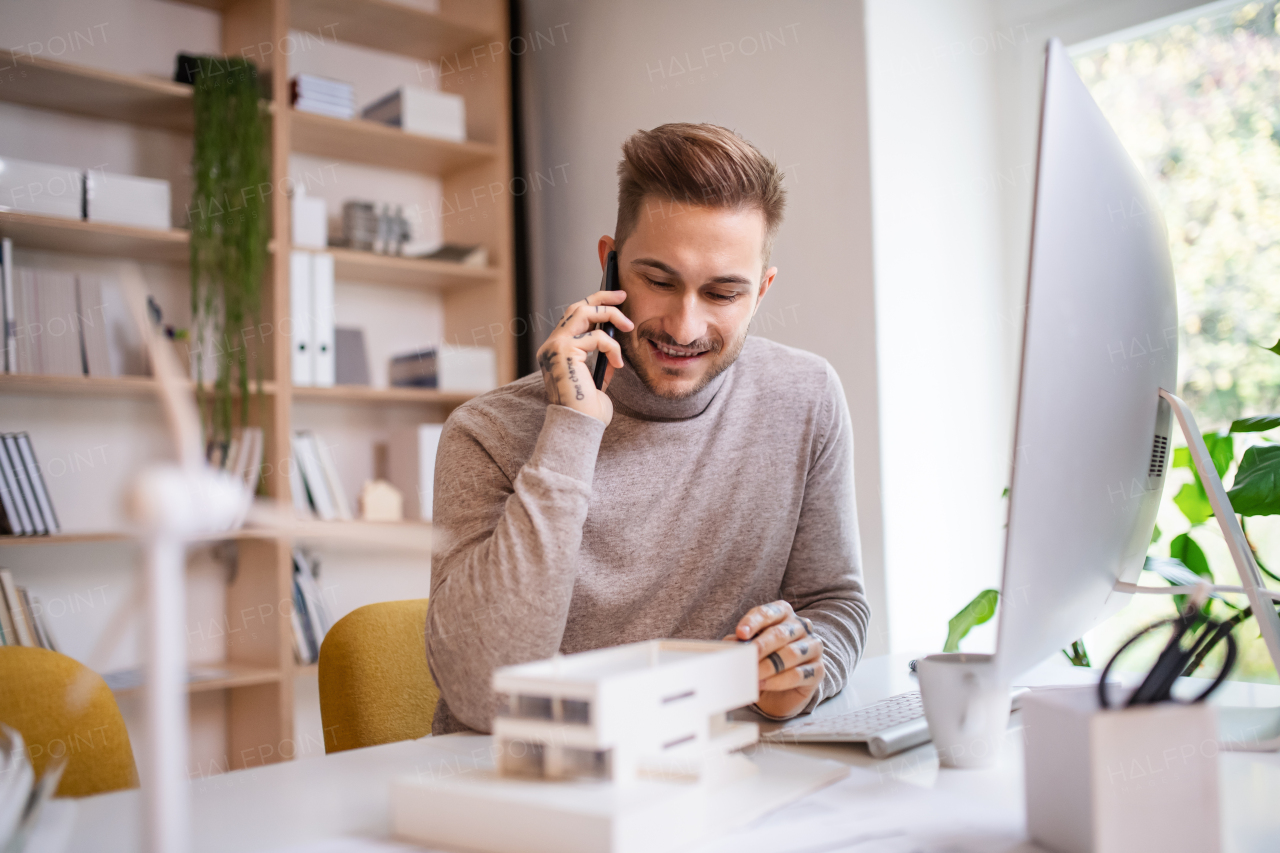 A young architect sitting at the desk indoors in office, using smartphone.