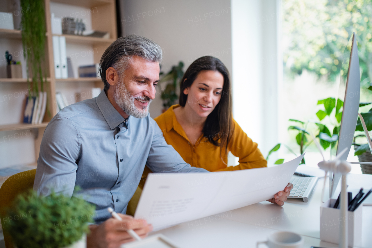 Mature architects sitting at the desk indoors in office, looking at blueprints.