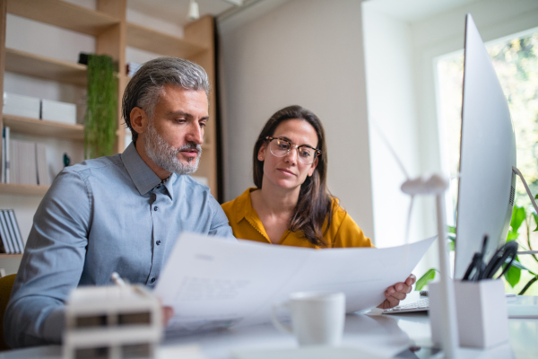 Mature architects sitting at the desk indoors in office, looking at blueprints.