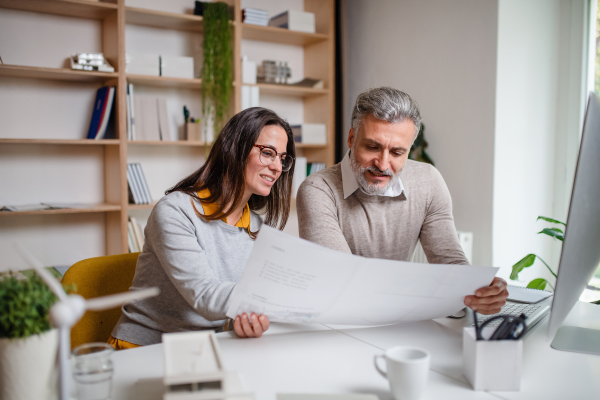 Mature architects sitting at the desk indoors in office, looking at blueprints.