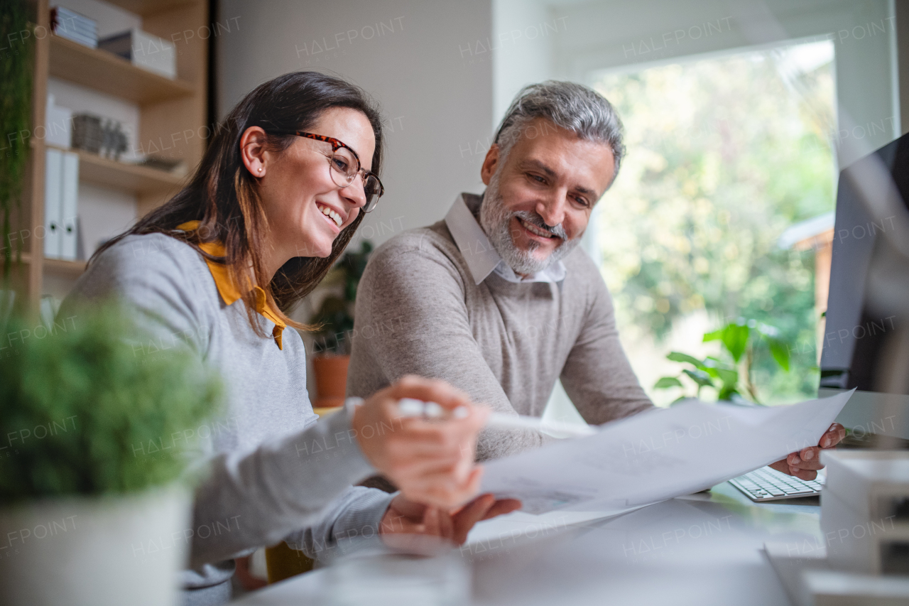 Mature architects sitting at the desk indoors in office, looking at blueprints.