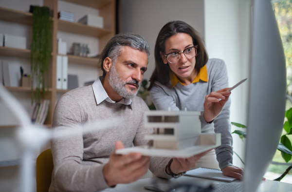 Mature architects with model of house sitting at the desk indoors in office, working.