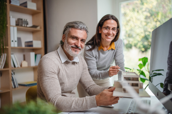 Mature architects with model of house sitting at the desk indoors in office, working.