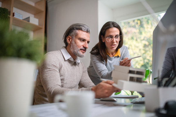 Mature architects with model of house sitting at the desk indoors in office, working.