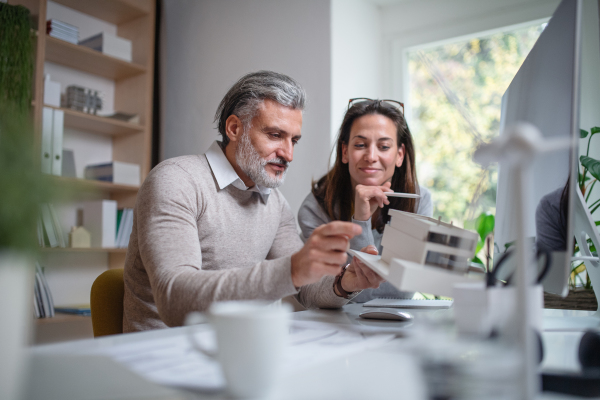 Mature architects with model of house sitting at the desk indoors in office, working.