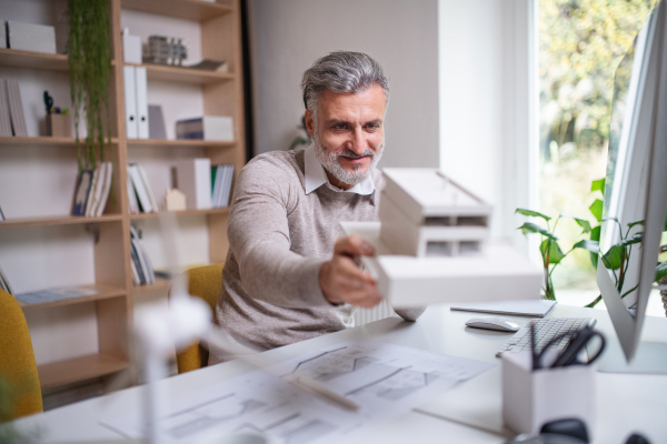 Mature architect with model of house sitting at the desk indoors in office, working.