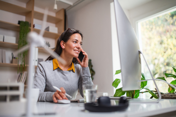 Attractive businesswoman sitting at the desk indoors in office, using smartphone and computer.