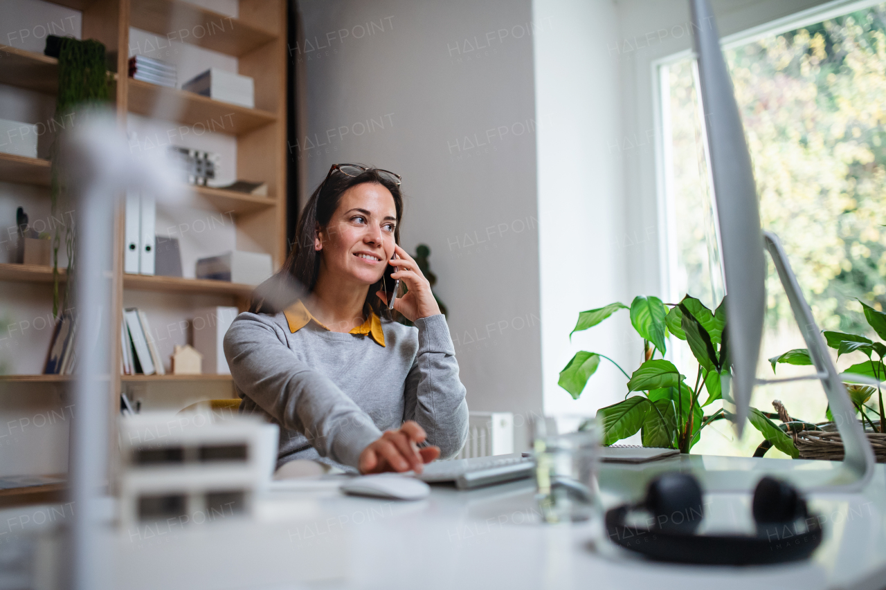 Attractive businesswoman sitting at the desk indoors in office, using smartphone and computer.