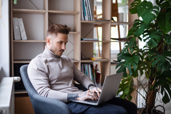 A young businessman sitting in armchair indoors in office, using laptop.