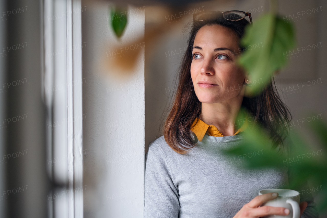 Attractive businesswoman with coffee standing by window indoors in office, resting.