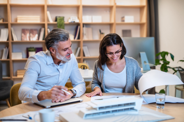 Mature architects sitting at the desk indoors in office, looking at blueprints.