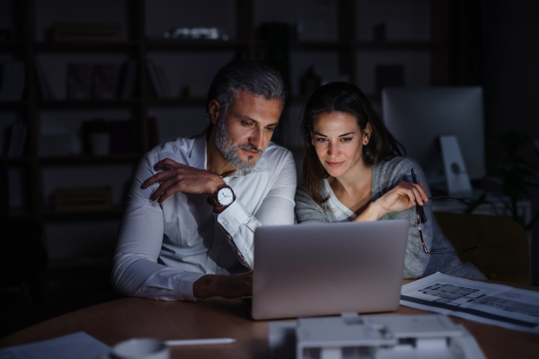 Mature architects with laptop sitting at the desk indoors in office, working late.