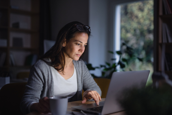 A businesswoman with laptop sitting at the desk indoors in dark office, working late.