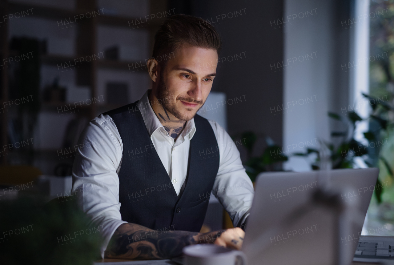 Young businessman with laptop sitting at the desk indoors in office, working late.