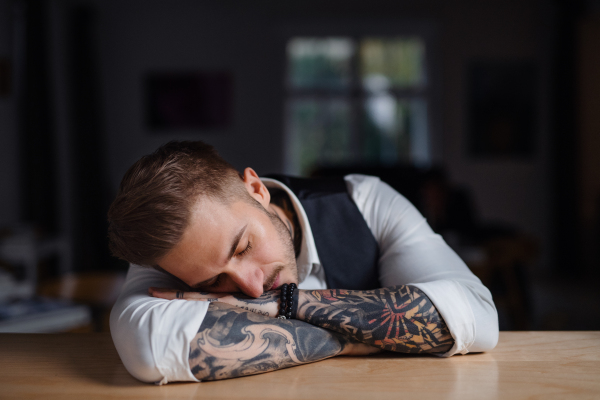 A tired young businessman sitting at the desk indoors in office, sleeping.