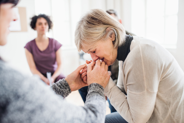 A senior depressed woman crying during group therapy, other people comforting her.