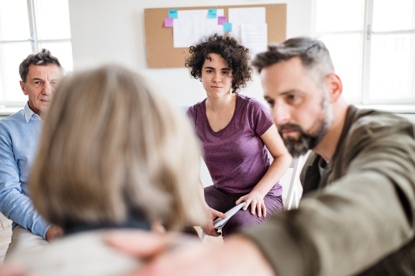 Serious men and women sitting in a circle during group therapy, supporting each other.