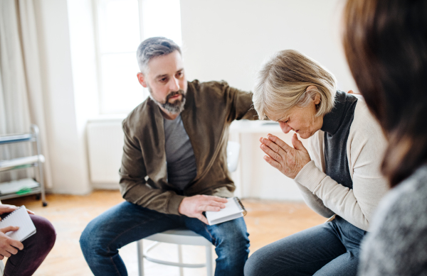 A senior depressed woman crying during group therapy, other people comforting her.