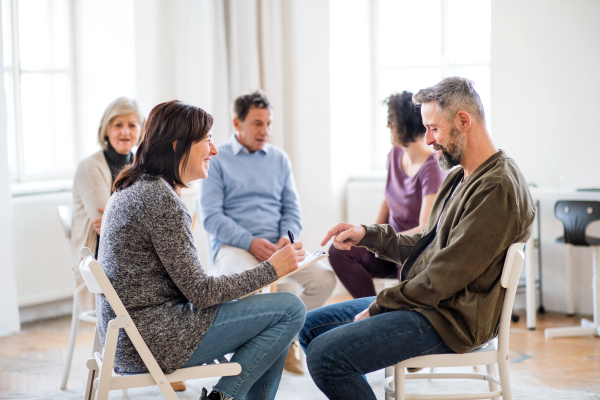 A senior counsellor with clipboard talking to a man during group therapy.