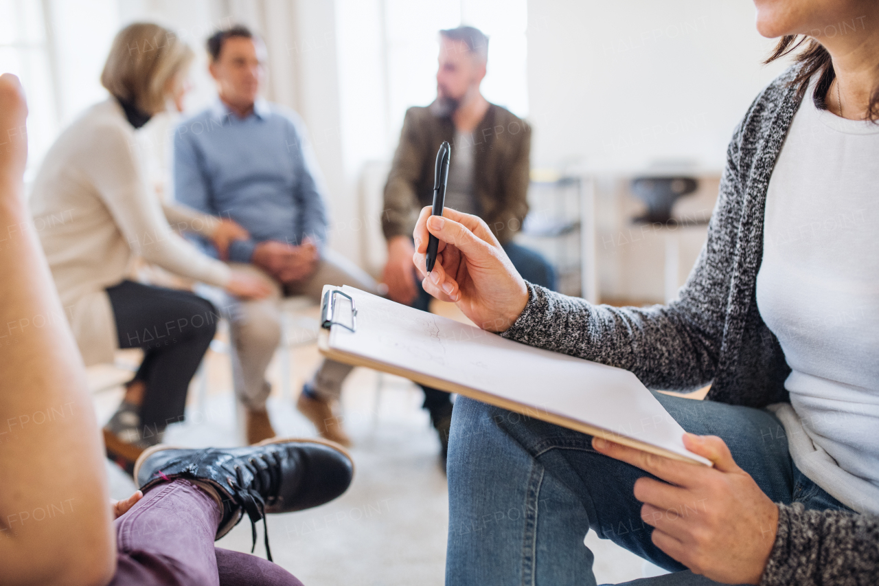 A midsection view of counsellor with clipboard talking to a client during group therapy.