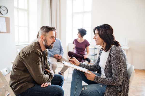 A senior counselor with clipboard talking to a man during group therapy.