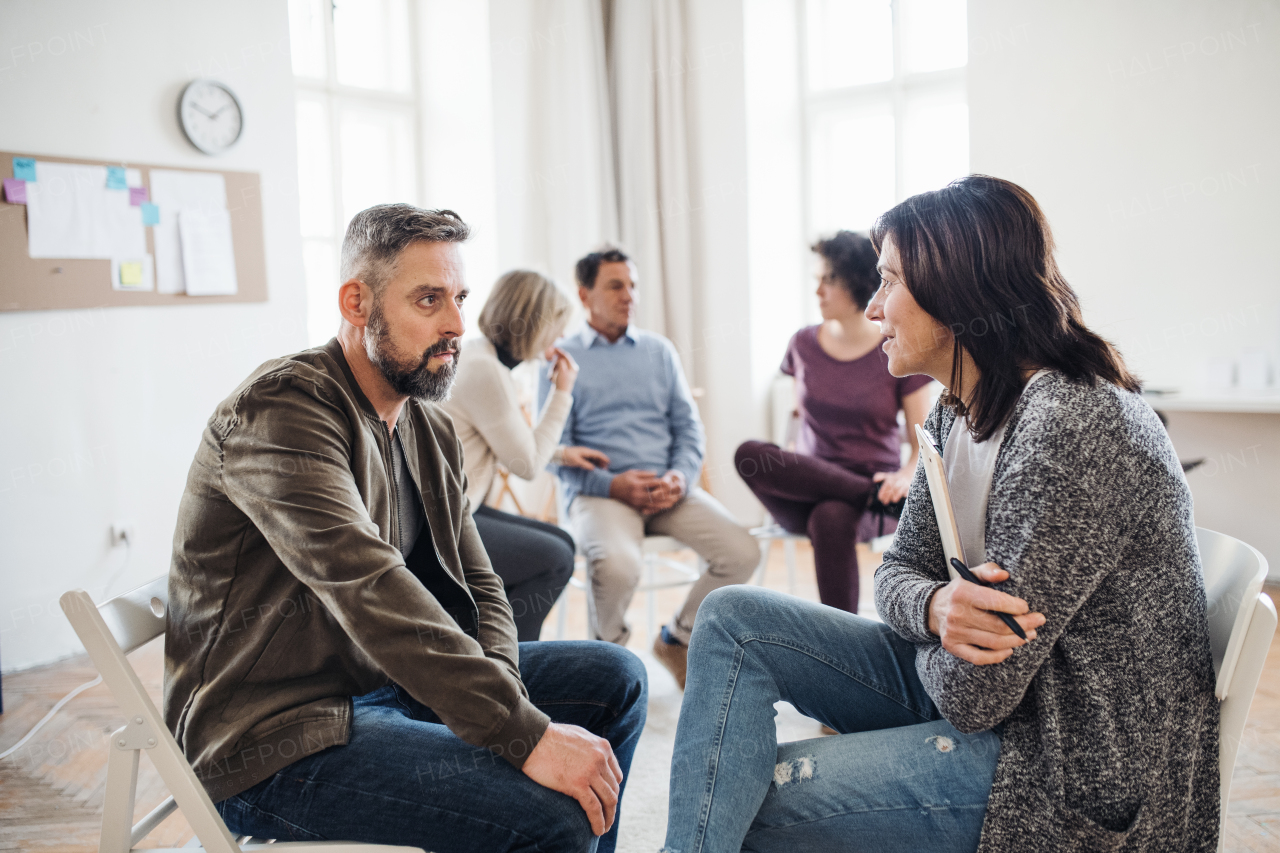 A senior counselor with clipboard talking to a man during group therapy.