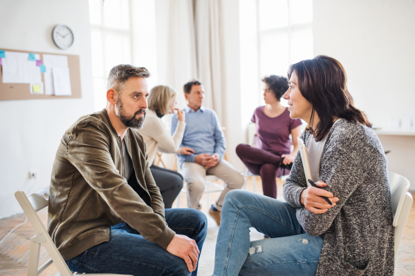 A senior counselor with clipboard talking to a man during group therapy.