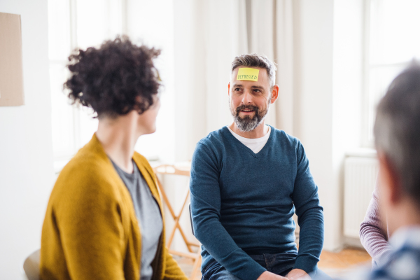 Men and women sitting in a circle during group therapy, adhesive notes with negative emotions on forehead.