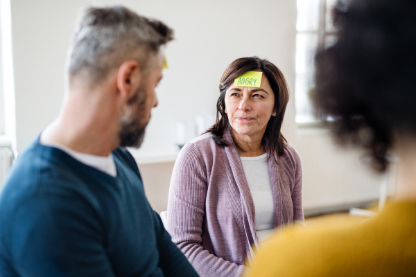 Men and women sitting in a circle during group therapy, adhesive notes with negative emotions on forehead.