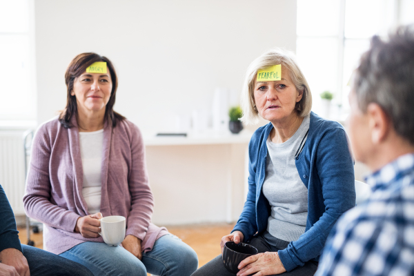 Men and women sitting in a circle during group therapy, adhesive notes with negative emotions on forehead.