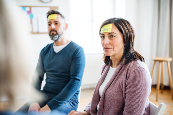 Men and women sitting in a circle during group therapy, adhesive notes with negative emotions on forehead.