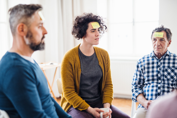 Men and women sitting in a circle during group therapy, adhesive notes with negative emotions on forehead.