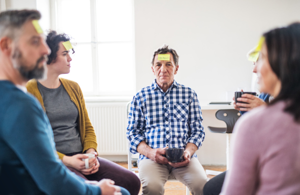 Men and women sitting in a circle during group therapy, adhesive notes with negative emotions on forehead.