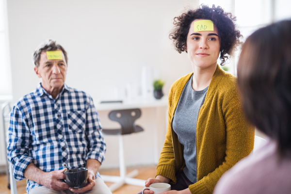 Men and women sitting in a circle during group therapy, adhesive notes with negative emotions on forehead.