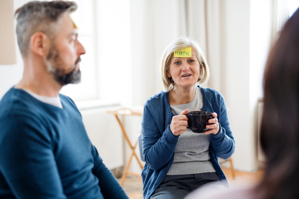 Men and women sitting in a circle during group therapy, adhesive notes with negative emotions on forehead.