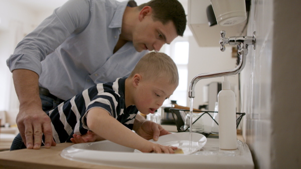 A father with down syndrome son indoors in kitchen, washing dishes.